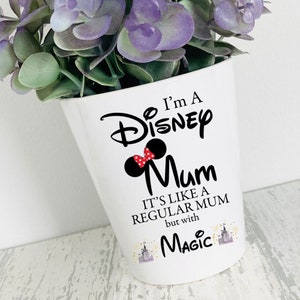 May include: White ceramic plant pot with the text "I'm A Disney Mum" and a Minnie Mouse design. The pot also says "It's like a regular mum but with Magic". Artificial purple and green flowers are in the pot.