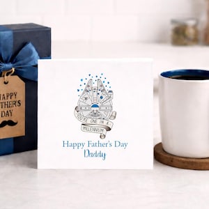 May include: A white greeting card featuring a line-art illustration of a spaceship, a banner with the text 'YOU'RE ONE IN A MILLENNIUM', and the printed text 'Happy Father's Day Daddy', positioned next to a dark blue gift box and a white mug.