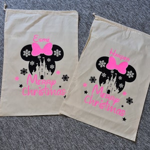 May include: Two white canvas gift bags with pink glitter Mickey Mouse ears and a pink bow. The bags have black glitter text that says "Merry Christmas" and the names "Esme" and "Harper".