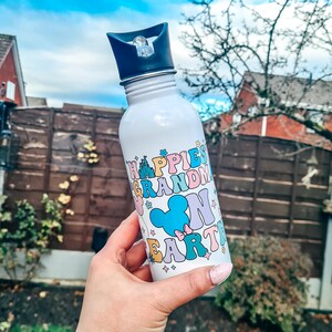 May include: White stainless steel water bottle with a black lid and a colourful design that says "Happiest Grandma on Earth" with a Mickey Mouse head and other Disney-themed graphics.