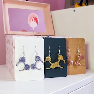 May include: Three pairs of dangle earrings shaped like mouse ears, each featuring a glittery finish. One pair is purple on a white card, one is gold on a black card, and one is pink on a brown card. Each earring has a silver-toned metal hook.