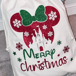 May include: White canvas Christmas gift bag with a red and green glitter design of Mickey Mouse's head with a bow and Cinderella's castle. The bag has the text "Merry Christmas" in green and red glitter.