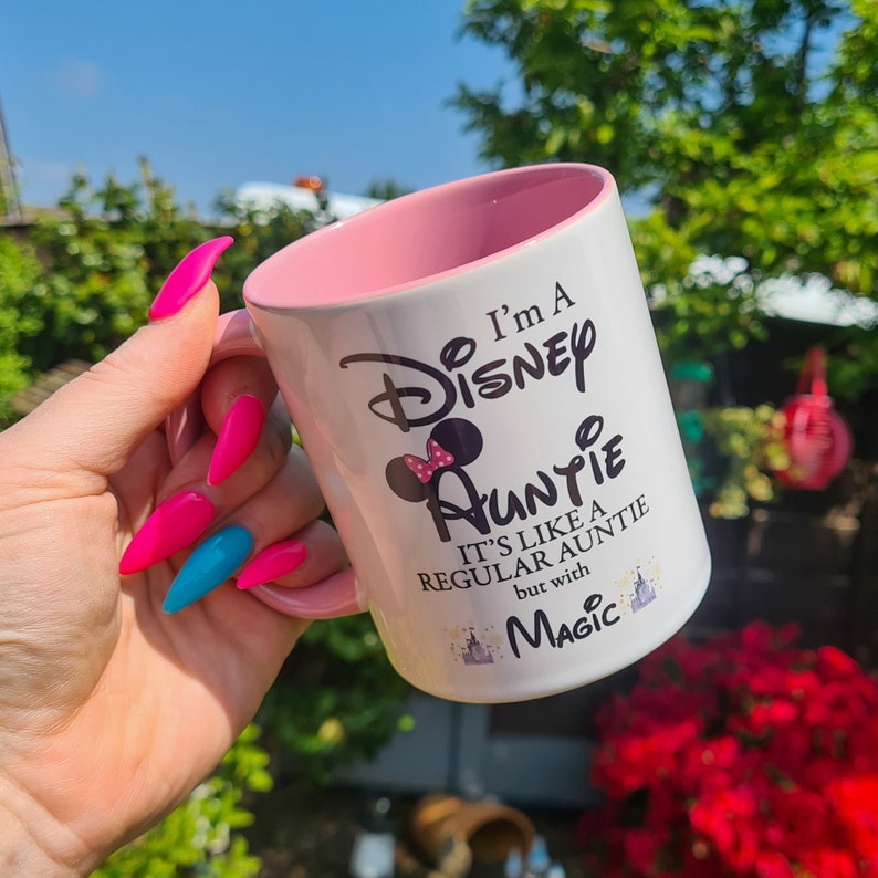 May include: White ceramic mug with a pink rim. The mug has a black graphic that says "I'm a Disney Auntie. It's like a regular auntie but with magic." The graphic also includes a black silhouette of Mickey Mouse's head with a pink bow.