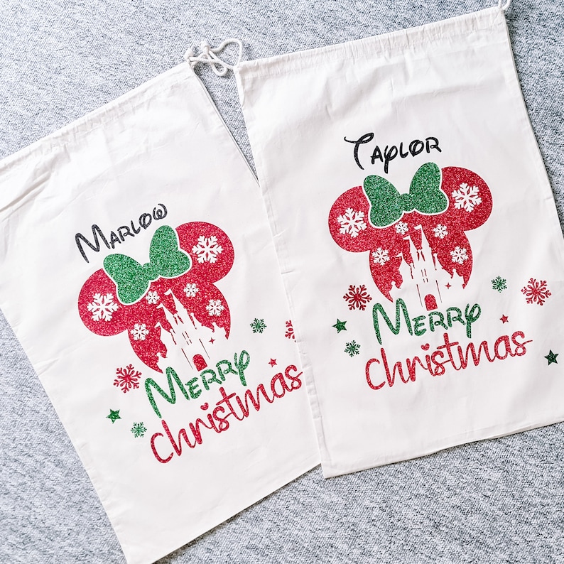 May include: Two white canvas gift bags with red and green glitter designs. The bags feature a Mickey Mouse silhouette with a castle and the text "Merry Christmas". One bag is personalized with the name "Marlow" and the other with the name "Taylor".