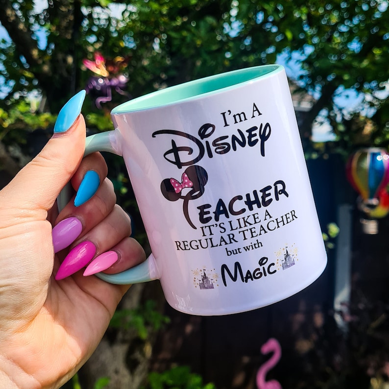 May include: White ceramic mug with a teal rim. The mug has a black graphic of Mickey Mouse ears with a pink bow and the text "I'm a Disney Teacher It's like a regular teacher but with Magic".