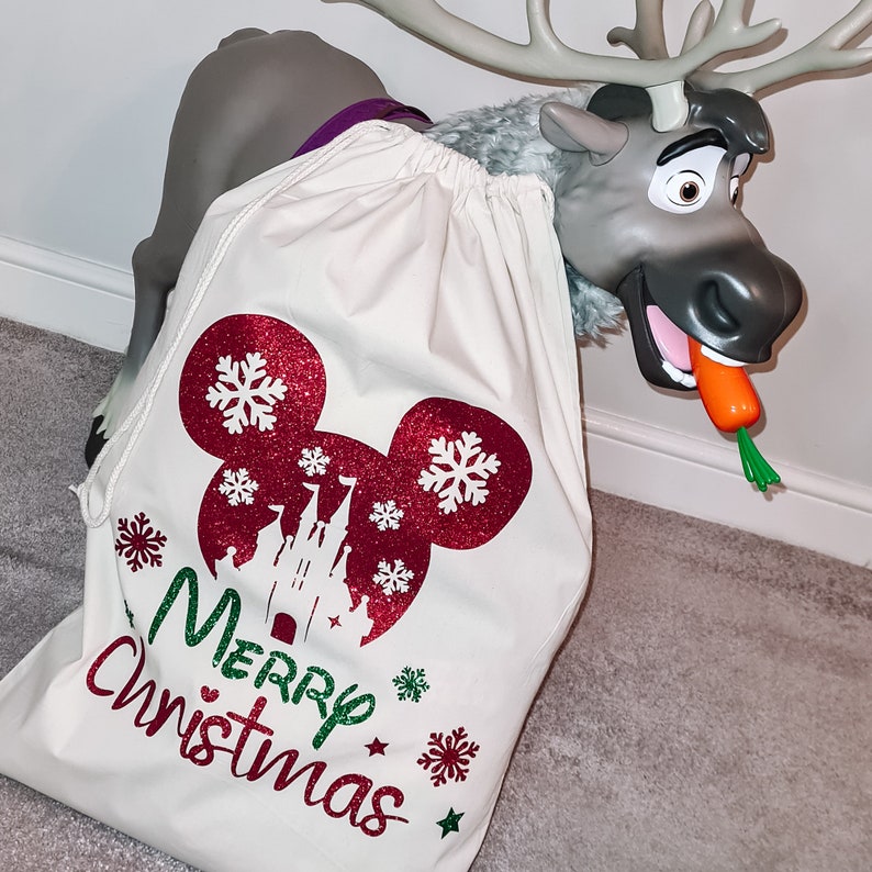 May include: A white canvas Christmas gift bag with a red glitter Mickey Mouse head and a red glitter castle design. The bag has green glitter snowflakes and the text "Merry Christmas" in green glitter.