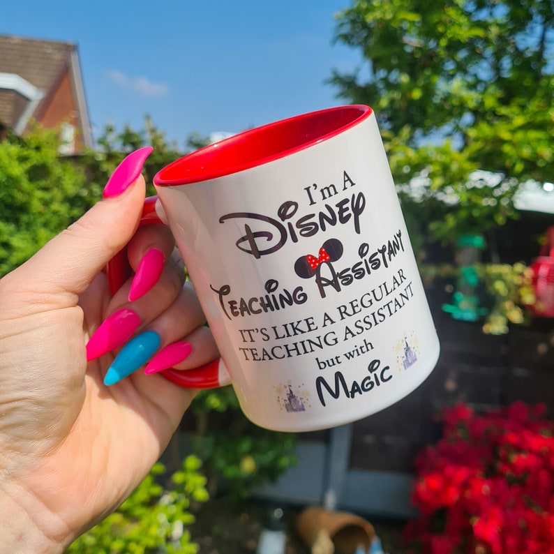 May include: White ceramic mug with a red rim. The mug has a black graphic that says "I'm a Disney Teaching Assistant. It's like a regular teaching assistant but with Magic." The graphic includes a black silhouette of Mickey Mouse's head with a red bow.