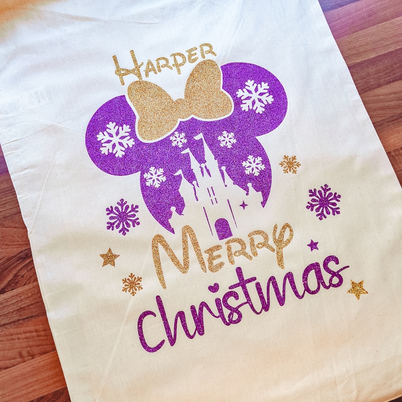 May include: A white canvas tote bag with a purple and gold glitter design. The design features Mickey Mouse ears with a bow, a castle, snowflakes, and the text "Harper Merry Christmas".