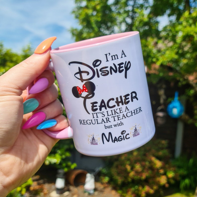 May include: White ceramic mug with a pink rim. The mug has a black graphic that says "I'm a Disney Teacher It's like a regular teacher but with Magic" with a red and black Mickey Mouse head with a red bow.