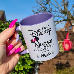 May include: White ceramic mug with a purple rim. The mug has a black and white graphic of Mickey Mouse with a pink bow and the text "I'm a Disney Nurse. It's like a regular nurse but with Magic."