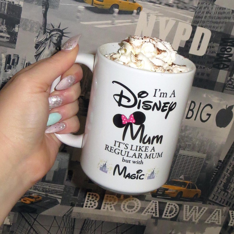 May include: White ceramic mug with a black and white graphic that says "I'm a Disney Mum. It's like a regular mum but with magic." The graphic also includes a pink bow and a black silhouette of Mickey Mouse's ears. The mug is filled with a hot beverage topped with whipped cream.