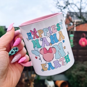 May include: A white ceramic mug with a pink rim and handle. The mug features a colourful graphic that says "Happiest Nana on Earth" with a cartoon Minnie Mouse head in the centre.