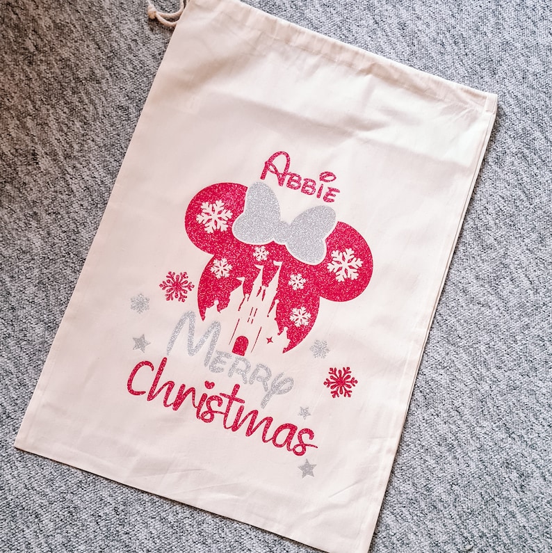 May include: A white canvas Christmas gift bag with a pink and silver glitter design featuring Mickey Mouse's head, a castle, snowflakes, and the text "Merry Christmas". The bag is personalized with the name "Abbie".