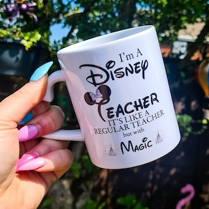 May include: White ceramic mug with a black design that says "I'm a Disney Teacher. It's like a regular teacher but with magic." The design also includes a black silhouette of Mickey Mouse's head with a pink bow.