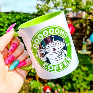 May include: White coffee mug with a green rim and a green logo featuring a cartoon alien holding a cup of coffee. The logo says "OoooOh! Coffee".