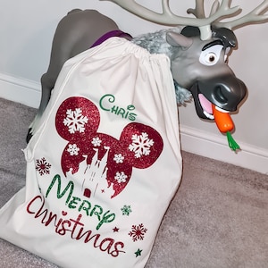May include: A white canvas Christmas gift bag with a red and white Mickey Mouse design, a green and white castle, and the text "Merry Christmas". The bag is personalized with the name "Chris".