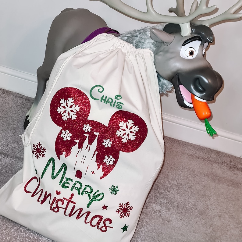 May include: A white canvas Christmas gift bag with a red and white Mickey Mouse design, a green and white castle, and the text "Merry Christmas". The bag is personalized with the name "Chris".