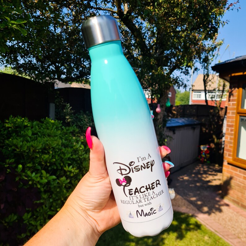May include: A turquoise and white stainless steel water bottle with a Disney design. The bottle has the text "I'm a Disney Teacher. It's like a regular teacher but with Magic."