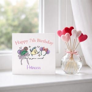 May include: A white square card with the text "Happy 7th Birthday" and the word "Princess". The card features illustrations of cartoon characters. A glass vase with heart-shaped decorations sits nearby.