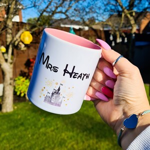 May include: White ceramic mug with a pink rim. The mug has a black and gold design of a castle with fireworks and the text "Mrs Heath".