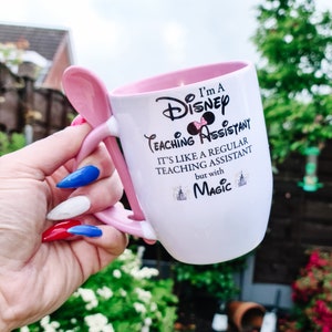 May include: White ceramic mug with a pink handle and rim. The mug has a black and white graphic that says "I'm a Disney Teaching Assistant. It's like a regular teaching assistant but with Magic." The graphic includes a black silhouette of Mickey Mouse with a pink bow.