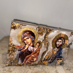 May include: A wooden plaque with a gold and white background features two religious icons. The left icon depicts the Virgin Mary holding a baby Jesus. The right icon depicts Jesus Christ.
