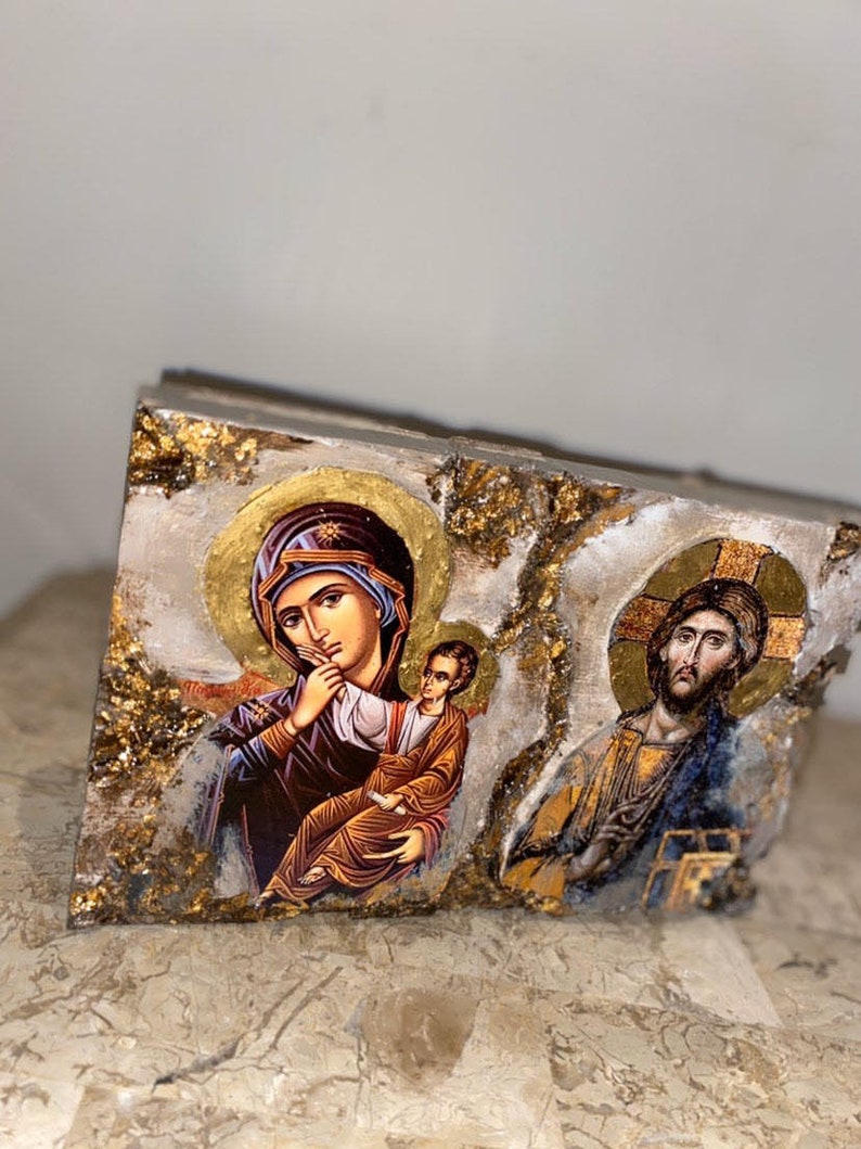 May include: A wooden plaque with a gold and white background features two religious icons. The left icon depicts the Virgin Mary holding a baby Jesus. The right icon depicts Jesus Christ.