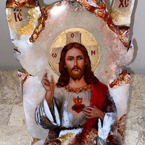Puede incluir: Un icono de madera pintado de Jesucristo con un corazón rojo y un halo dorado. El icono está en un soporte y tiene un fondo blanco desgastado con detalles dorados.