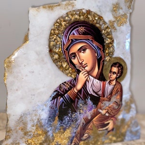 Puede incluir: Una placa de mármol blanco con detalles dorados presenta un icono religioso de la Virgen María y el niño Jesús. El icono está pintado en un estilo tradicional con colores vibrantes y detalles intrincados.