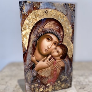May include: A hand-painted icon depicting the Virgin Mary holding the baby Jesus. The icon is painted on a wooden panel with gold leaf accents. The background is a dark brown with hints of gold and silver.
