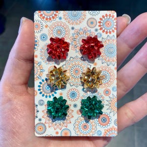 Könnte beinhalten: Ein Set aus sechs schleifenförmigen Ohrringen in Rot, Gold und Grün. Die Ohrringe sind auf einer dekorativen Karte mit einem Blumenmuster in Blau, Orange und Weiß präsentiert. Die Ohrringe sind klein und glänzend.
