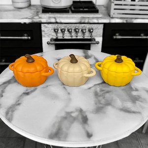 Può includere: Tre ciotole in ceramica a forma di zucca in miniatura, arancioni, crema e gialle. Ogni ciotola ha un gambo marrone e piccole maniglie. Le ciotole sono disposte su una superficie rotonda con motivo marmorizzato.