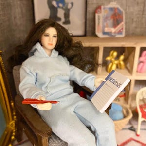 Pu&ograve; includere: Una bambola con una felpa blu chiaro e pantaloni abbinati &egrave; seduta su una sedia di legno. La bambola tiene una penna rossa e un quaderno blu con la scritta "NOTEBOOK" sulla copertina.