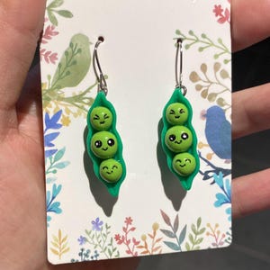 Puede incluir: Un par de pendientes caprichosos de vainas de guisantes. Cada pendiente presenta tres guisantes verdes sonrientes dentro de una vaina verde. Los pendientes cuelgan de alambres plateados y se muestran en una tarjeta decorativa.