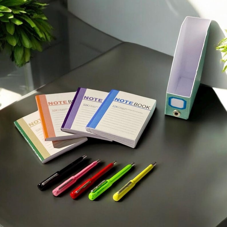 Pu&ograve; includere: Quattro piccoli quaderni con la scritta "NOTE BOOK" sulla copertina, impilati su una superficie grigia. I quaderni sono di diversi colori: arancione, viola, blu e verde. Cinque penne di diversi colori: nero, rosa, rosso, verde e giallo sono anche sulla superficie.