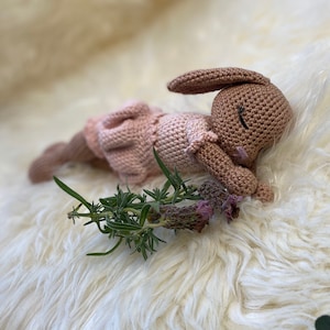 Könnte beinhalten: Eine gehäkelte rosa und braune Hasenpuppe liegt auf einer weißen flauschigen Oberfläche mit grünen Lavendelzweigen.