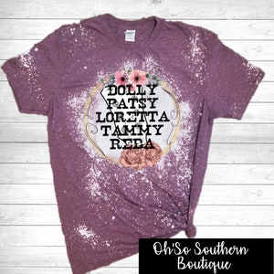 May include: A mauve t-shirt with a bleached design. The shirt features a floral graphic with the names Dolly, Patsy, Loretta, Tammy, and Reba. The bottom right corner has the text "Oh So Southern Boutique".