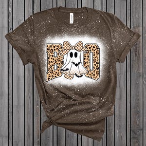 May include: Brown t-shirt with a white splattered design. The shirt features a graphic that says "BOO" in leopard print letters with a cartoon ghost wearing a bow. The ghost has a checkered pattern on its body.
