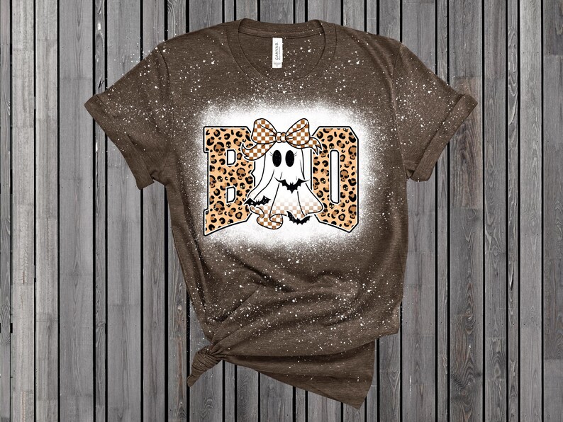 May include: Brown t-shirt with a white splattered design. The shirt features a graphic that says "BOO" in leopard print letters with a cartoon ghost wearing a bow. The ghost has a checkered pattern on its body.