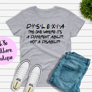 May include: A gray t-shirt with the word "DYSLEXIA" in colorful letters, followed by the text "THE ONE WHERE IT'S A DIFFERENT ABILITY, NOT A DISABILITY." The shirt is paired with blue jeans and white sneakers.