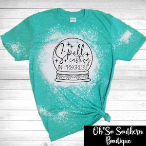 May include: A turquoise t-shirt with a bleached design and the words "Spell Casting In Progress" inside a crystal ball graphic. The shirt has a crew neck and short sleeves. The bottom right corner of the image has the text "Oh'So Southern Boutique".