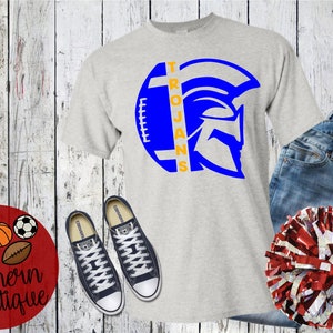 May include: A light gray t-shirt with a blue and orange Trojan helmet design, paired with blue jeans, black sneakers, and a red and white pom-pom. The shirt features the word "Trojans". A red circle with the text "Oh So Southern Boutique" is also visible.