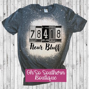 May include: A dark gray t-shirt with a bleach-splattered design featuring a vintage-style flip-style sign with the numbers 78418 and the text "Flour Bluff". The text "Oh So Southern Boutique" is printed in pink at the bottom of the image.