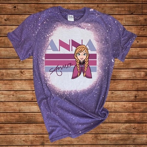 May include: A purple t-shirt with a bleached effect. The design includes the name "ANNA" in a stylized font, a character illustration with blonde braids, and a signature. The t-shirt is made of a soft material.
