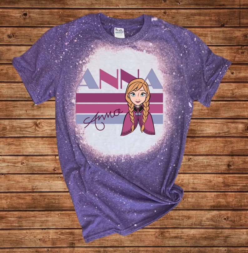 May include: A purple t-shirt with a bleached effect. The design includes the name "ANNA" in a stylized font, a character illustration with blonde braids, and a signature. The t-shirt is made of a soft material.