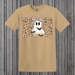May include: Tan t-shirt with a graphic that says "BOO". The letters are leopard print with a white ghost wearing a checkered bow. The ghost has black eyes and a checkered bottom. The shirt is on a wooden background.