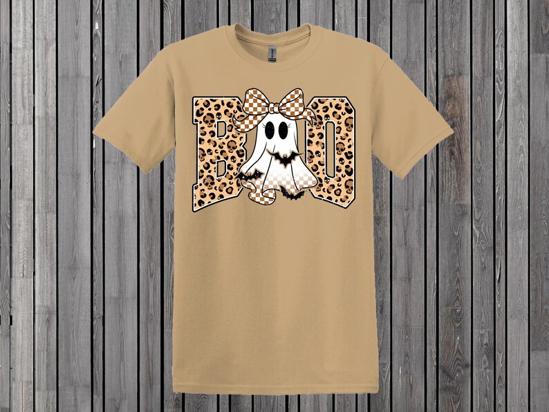 May include: Tan t-shirt with a graphic that says "BOO". The letters are leopard print with a white ghost wearing a checkered bow. The ghost has black eyes and a checkered bottom. The shirt is on a wooden background.
