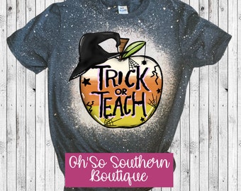 May include: A gray t-shirt with a distressed design featuring a graphic of an apple with a witch hat and the words "Trick or Teach". The apple design incorporates candy corn colors and spiderwebs, perfect for Halloween or teacher appreciation.