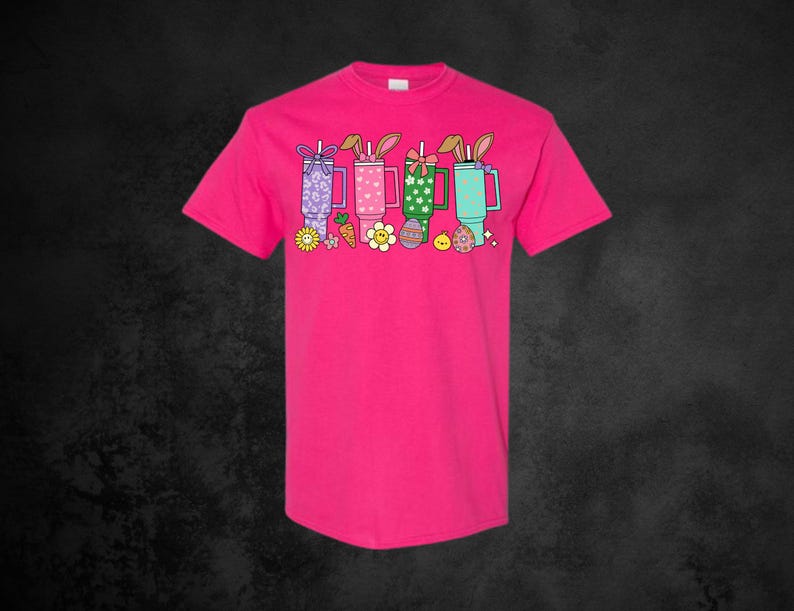 May include: A bright pink t-shirt featuring a colorful graphic of four Easter-themed drinks with bunny ears, flowers, and carrots. The design includes pastel colors and a playful aesthetic, perfect for the spring season.