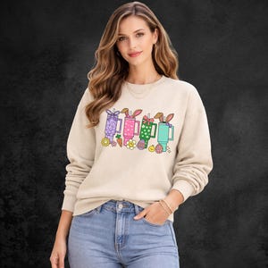 May include: Beige sweatshirt featuring a colorful graphic of Easter-themed mugs with bunny ears, carrots, and flowers. The design includes purple, pink, green, and blue mugs. The sweatshirt is worn by a person in blue jeans.